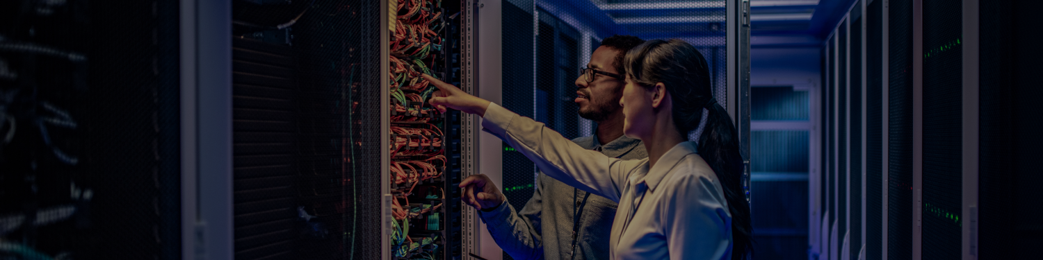 standing in a server room, interacting with network equipment. The individual appears to be working on a complex array of colorful wired connections within a server rack. The environment suggests a focus on technology and data management, with an emphasis on the maintenance or setup of network infrastructure. Mission Control NOC and Helpdesk, NOC, Helpdesk, IT Services, MSP Services, Outsourced MSP.